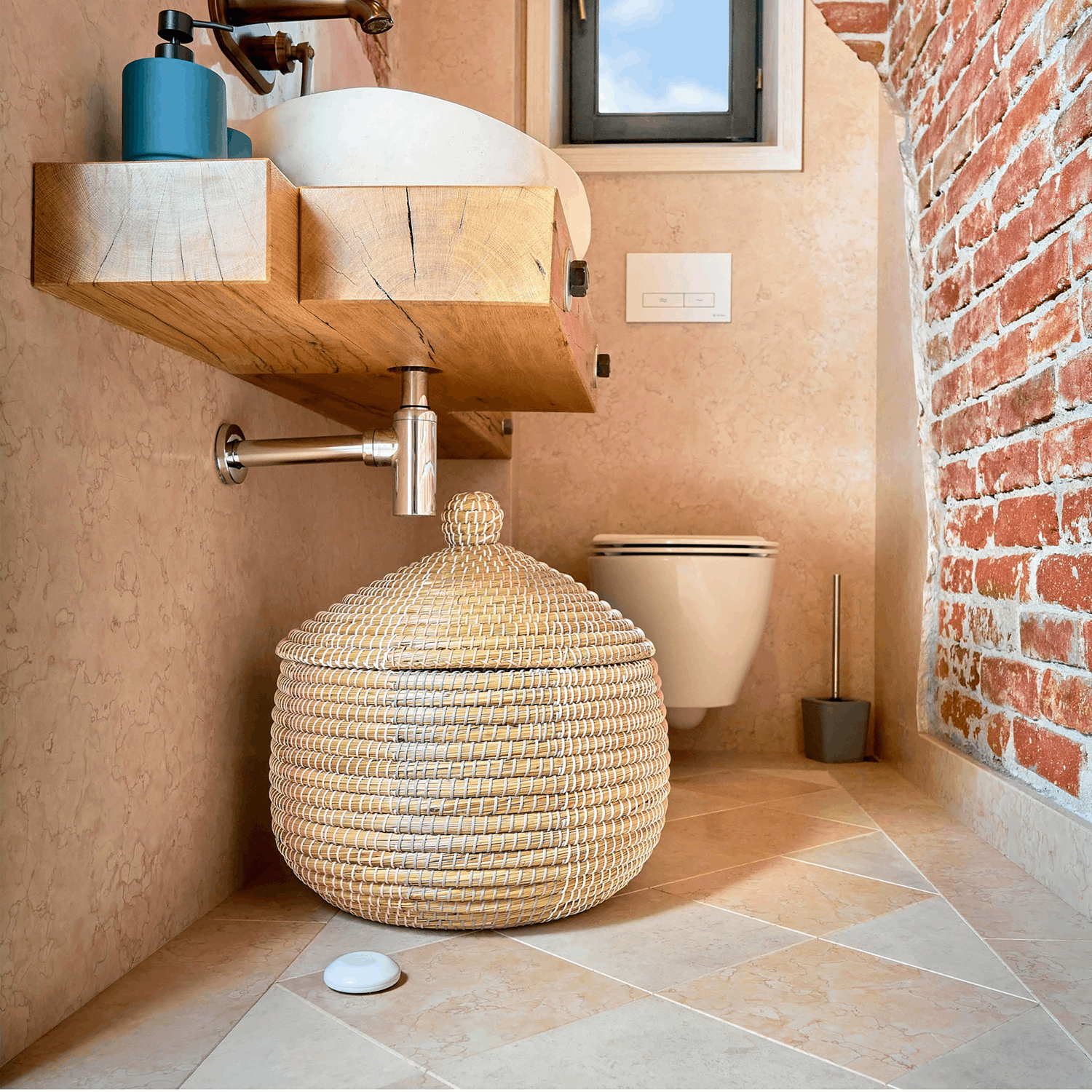 Interior de baño. Una cesta tejida, un lavabo de madera y un inodoro. Paredes beige y de ladrillo rojo.