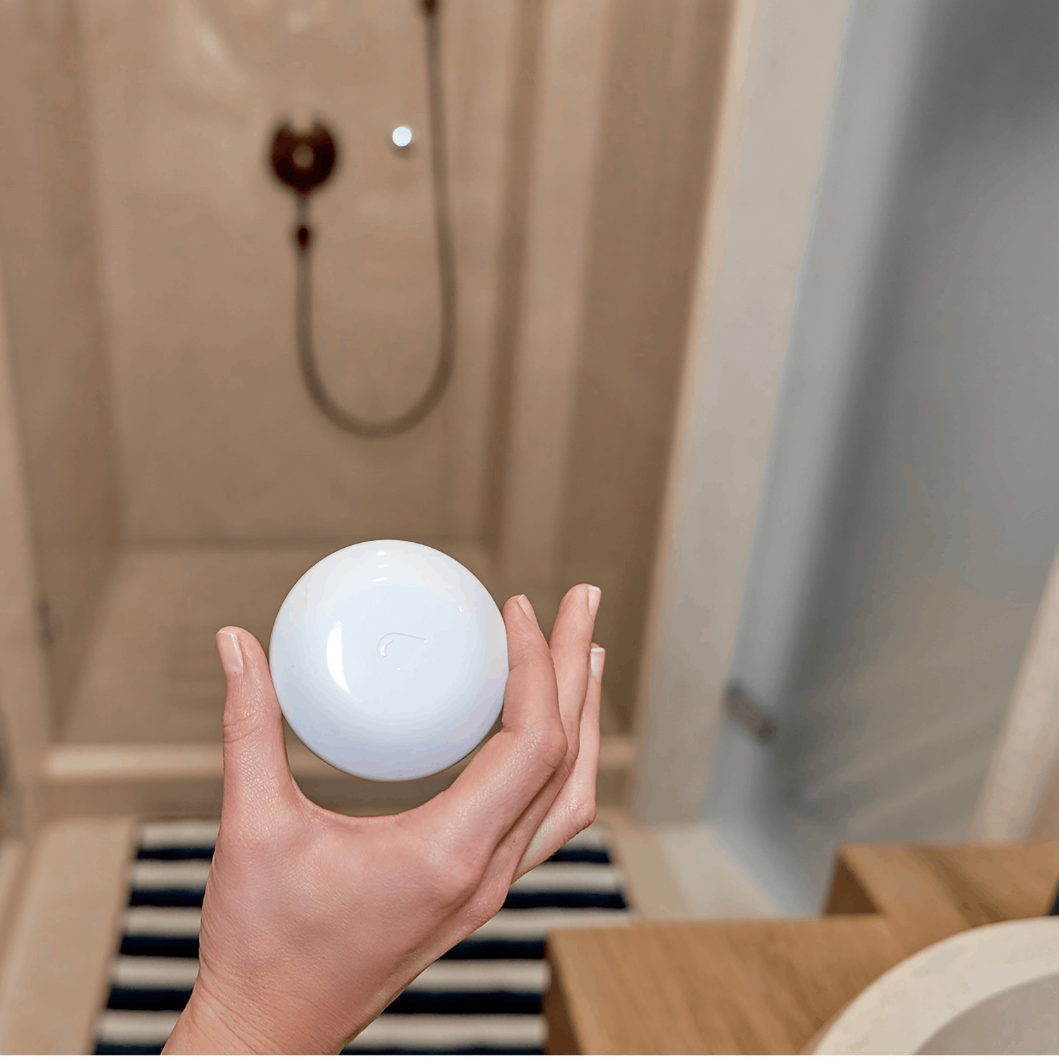 Una mano sosteniendo un objeto blanco y redondo en un baño con una ducha en el fondo.