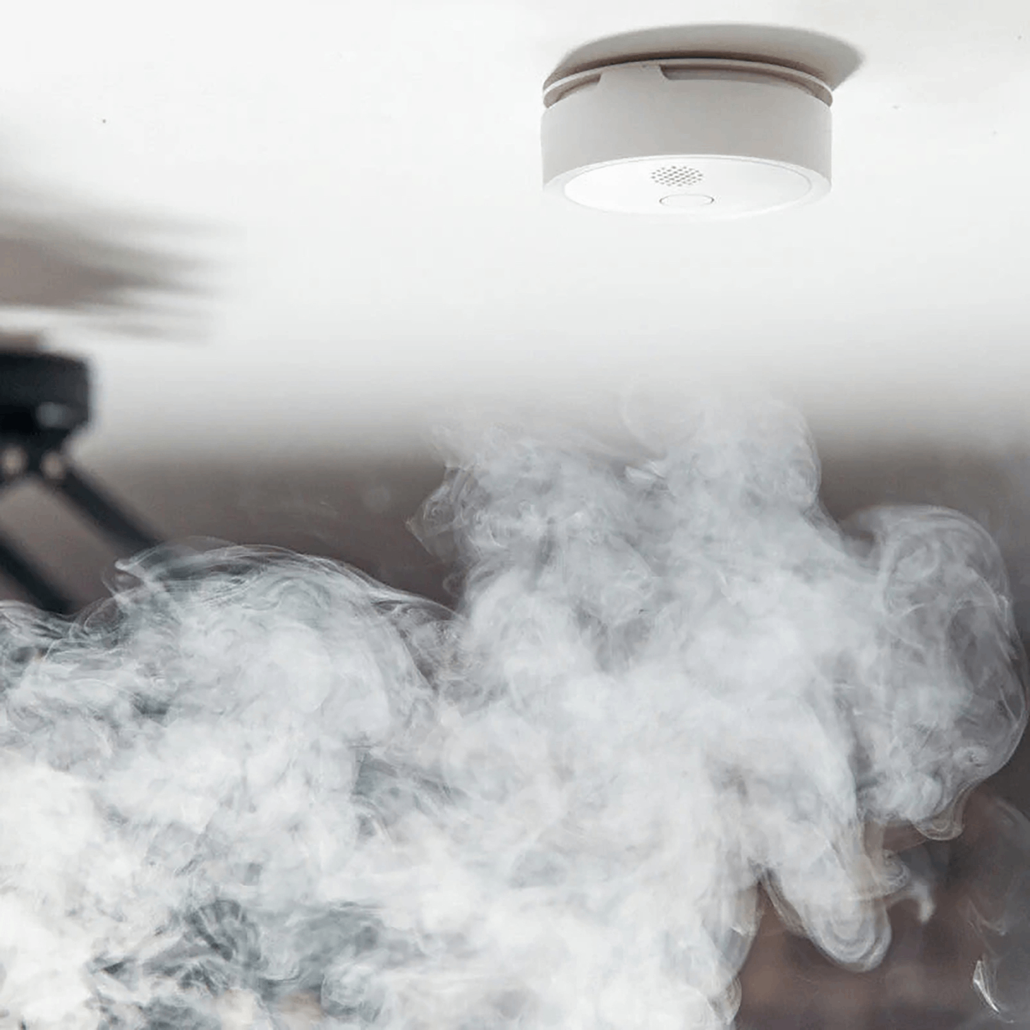 Detector de humo en un techo blanco con humo ondulante debajo, creando una nube gris.