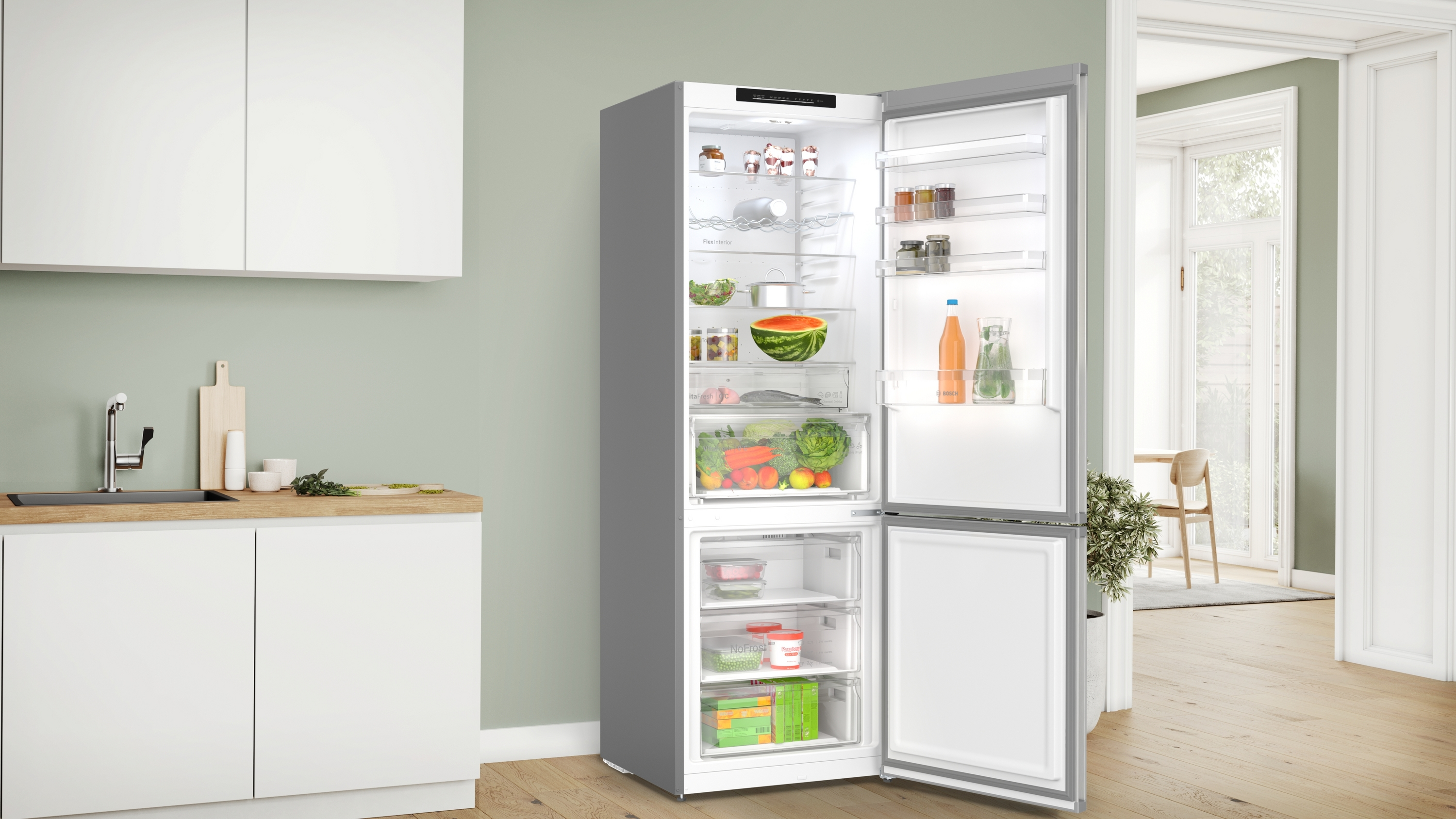 Ein offener Kühlschrank in einer Küche. Im Inneren befinden sich Früchte, Gemüse und Getränke. Die Küche hat weiße Schränke und ein Waschbecken.