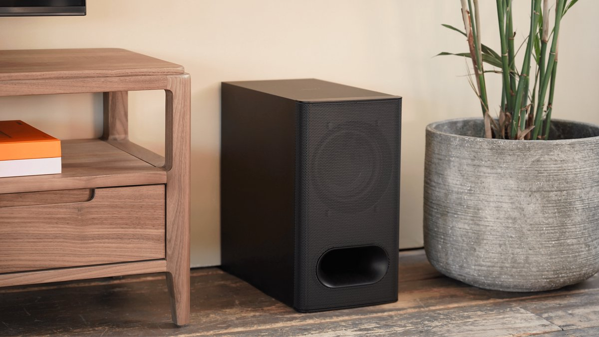 Czarny subwoofer obok rośliny w doniczce. Brązowy drewniany stół.