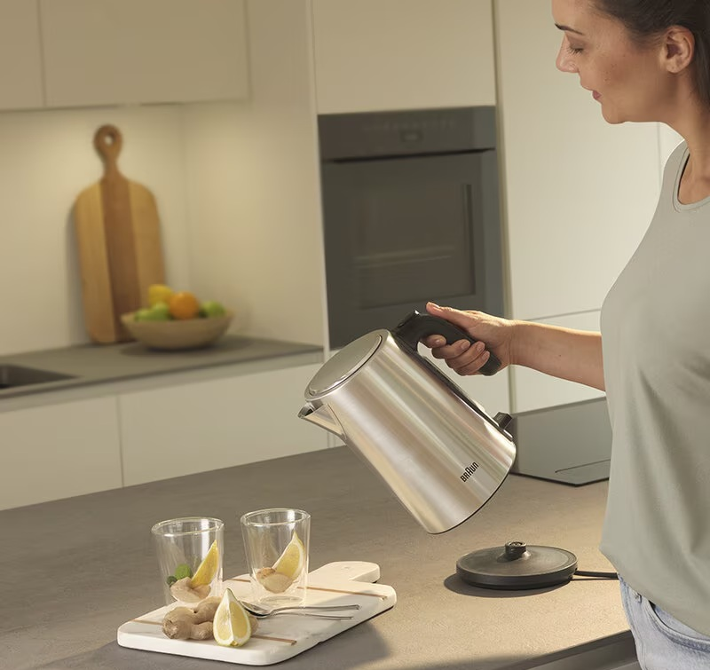 Kobieta nalewa wodę ze srebrnego czajnika do szklanki, z cytrynami i imbirem na blacie w nowoczesnej kuchni.