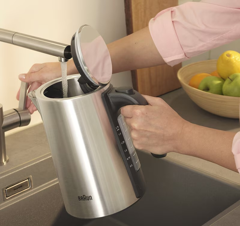 Osoba napełnia srebrny czajnik wodą z kranu. W tle stoi misa z owocami.