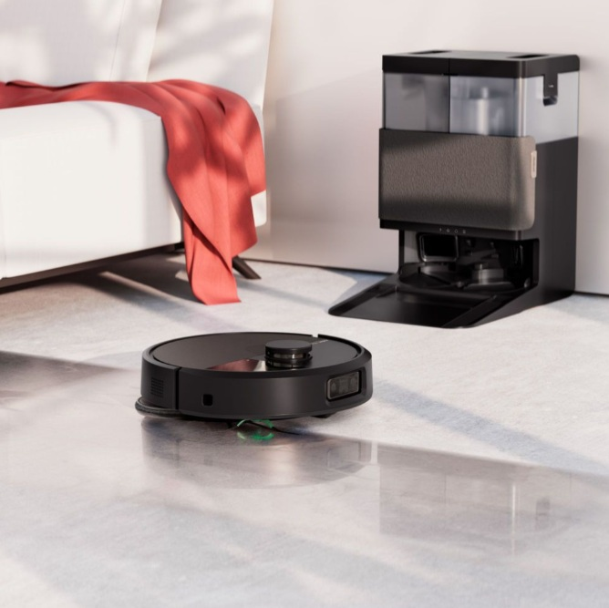 Czarny robot odkurzający na błyszczącej podłodze, obok stacji ładowania i czyszczenia.