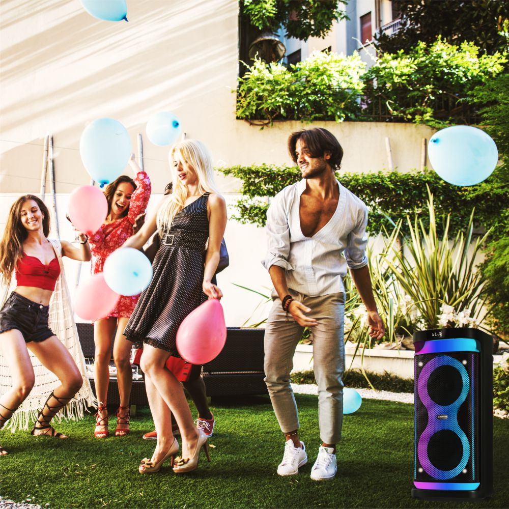 Ludzie tańczą na imprezie. Ludzie trzymają balony. Głośnik znajduje się po prawej stronie. Drzewa i budynek w tle.