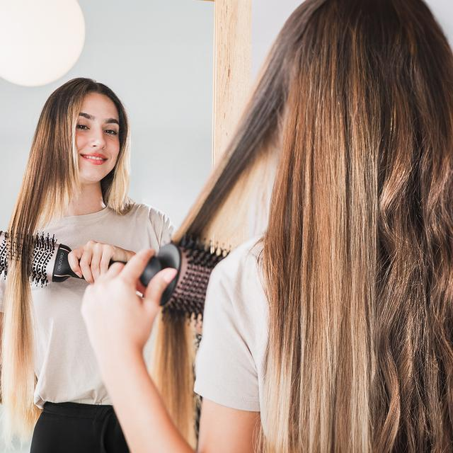 Kobieta używająca okrągłej szczotki na swoich długich, jasnobrązowych włosach, w lustrze.