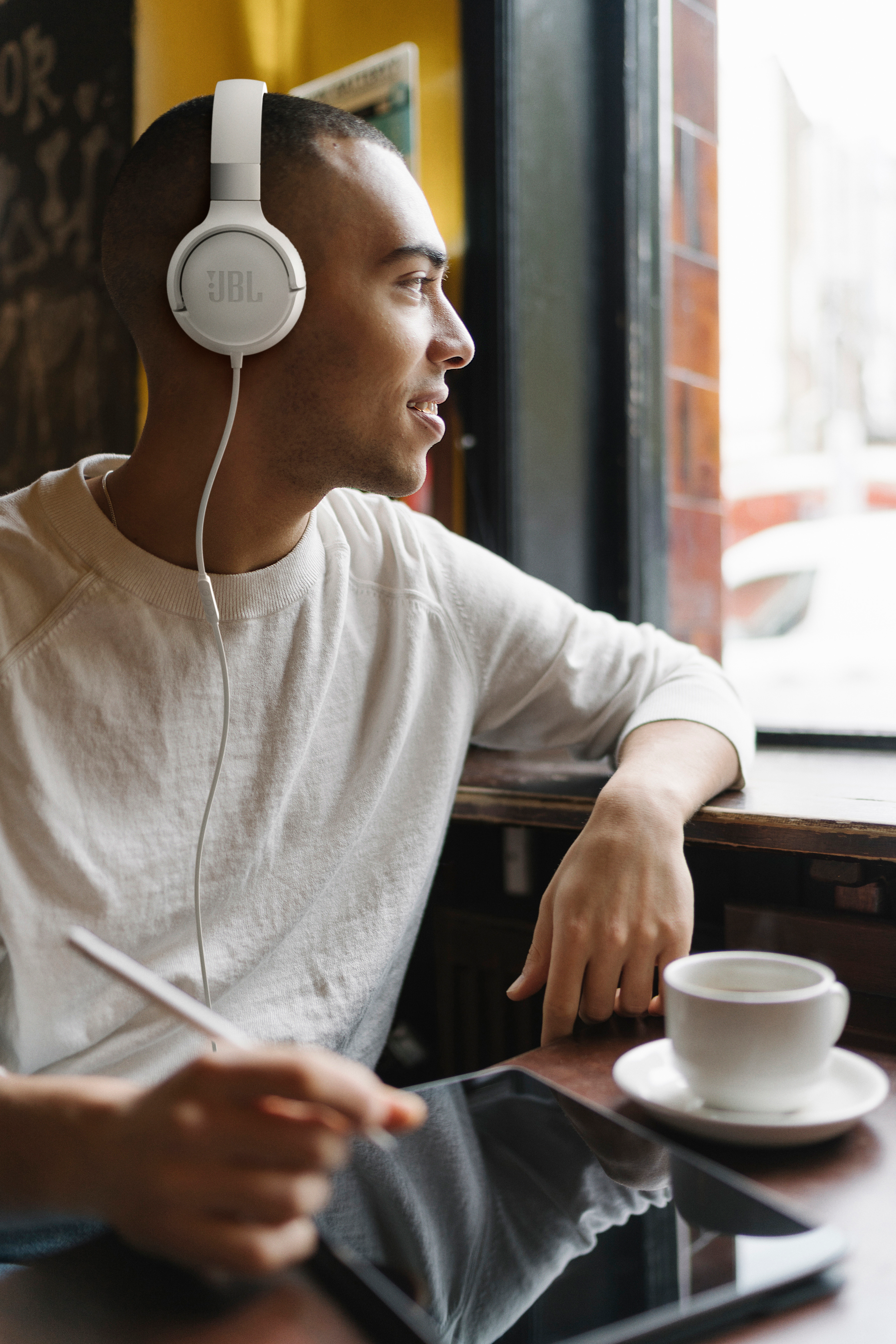 Mann mit weißen JBL-Kopfhörern, schaut aus dem Fenster. Er hat ein Tablet und eine Tasse Kaffee auf dem Tisch vor sich.