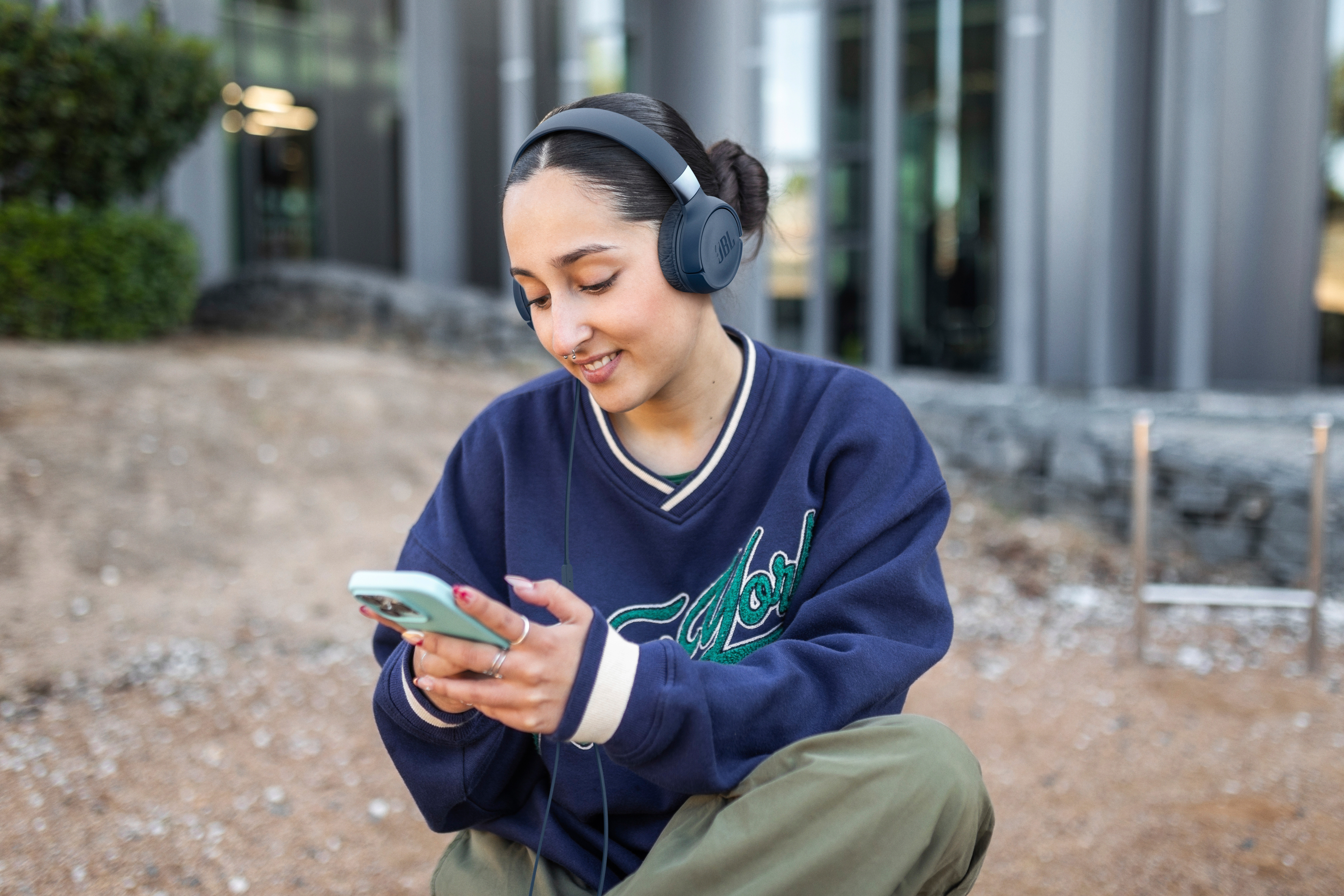 Eine Frau mit Kopfhörern schaut lächelnd auf ihr Handy. Sie trägt einen blauen Pullover und khakifarbene Hose und sitzt draußen. Gebäude im Hintergrund.