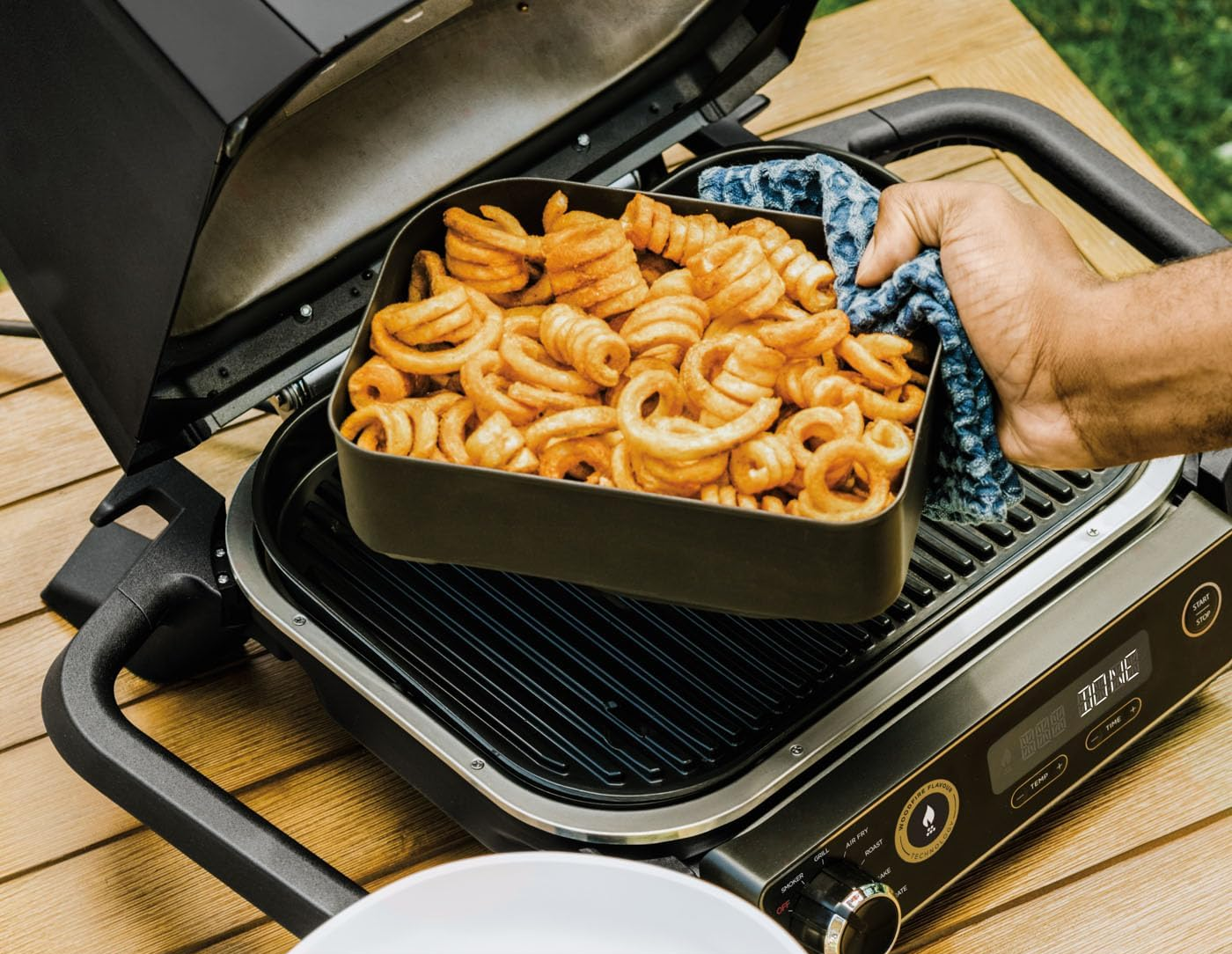 Személy egy tál göndör sült krumplit vesz le a grillről. Kültéri asztali környezet.