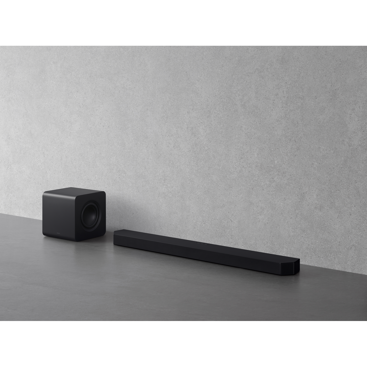 Black soundbar and subwoofer on gray floor, against gray wall. Modern home audio setup.