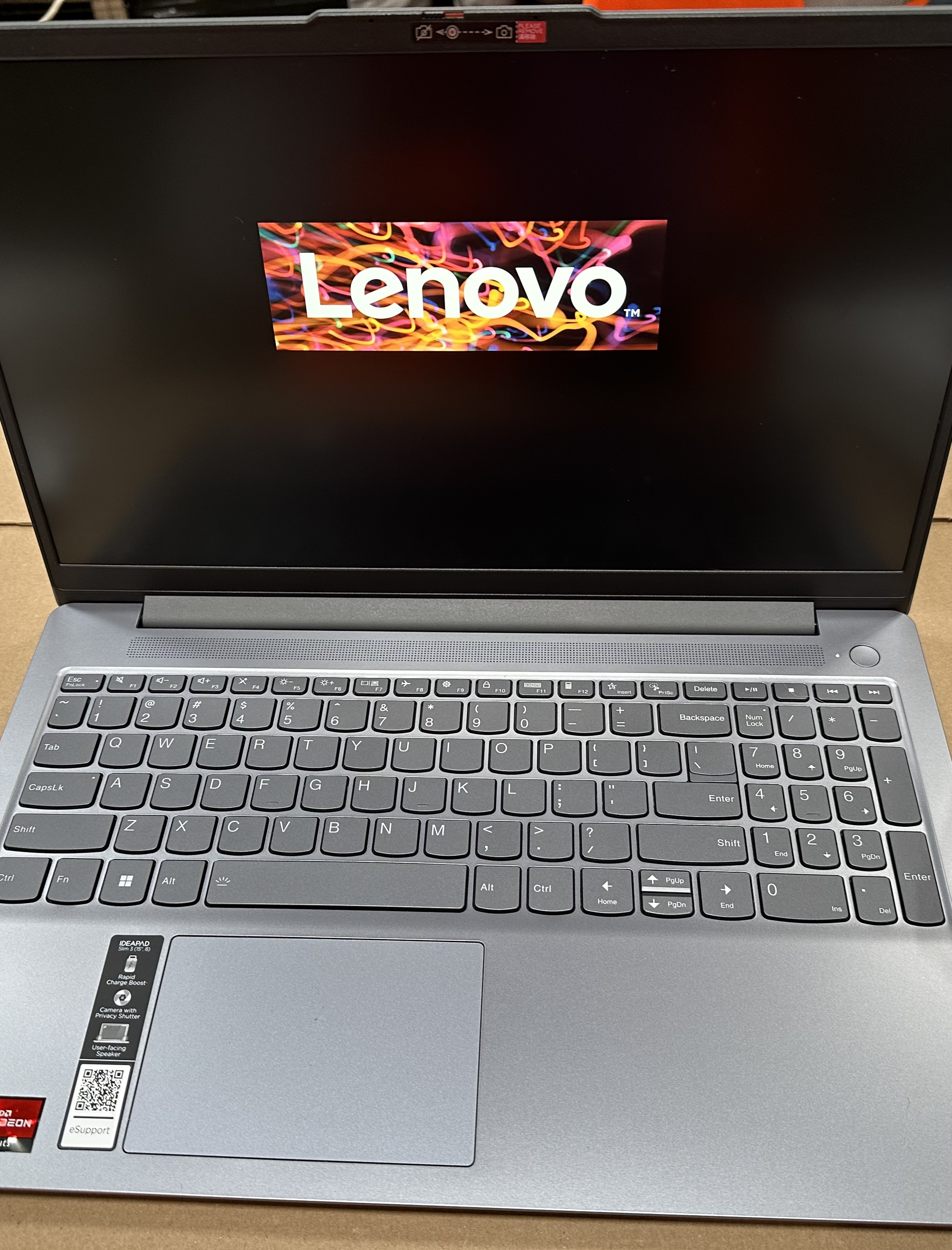 Laptop z logo Lenovo na ekranie, klawiatura widoczna poniżej. Laptop jest otwarty.