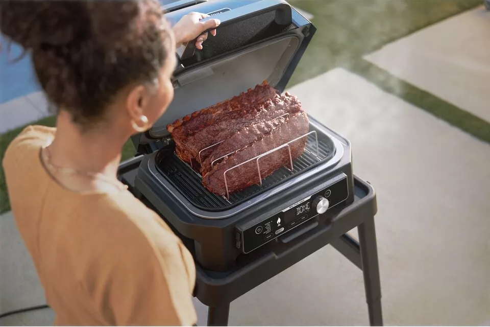 Nő bordákat süt egy kültéri grillen. A grill egy fekete állványon van.