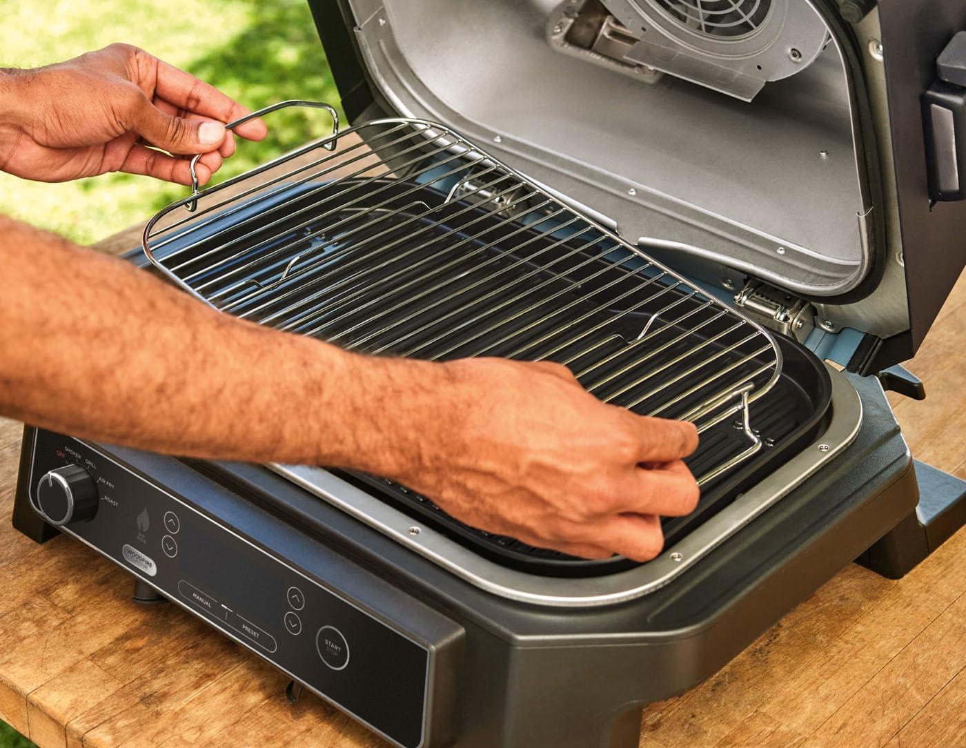 Kezek helyezik a grillrácsot egy fekete kültéri grillbe. A grill fa felületen van, zöld háttérrel.
