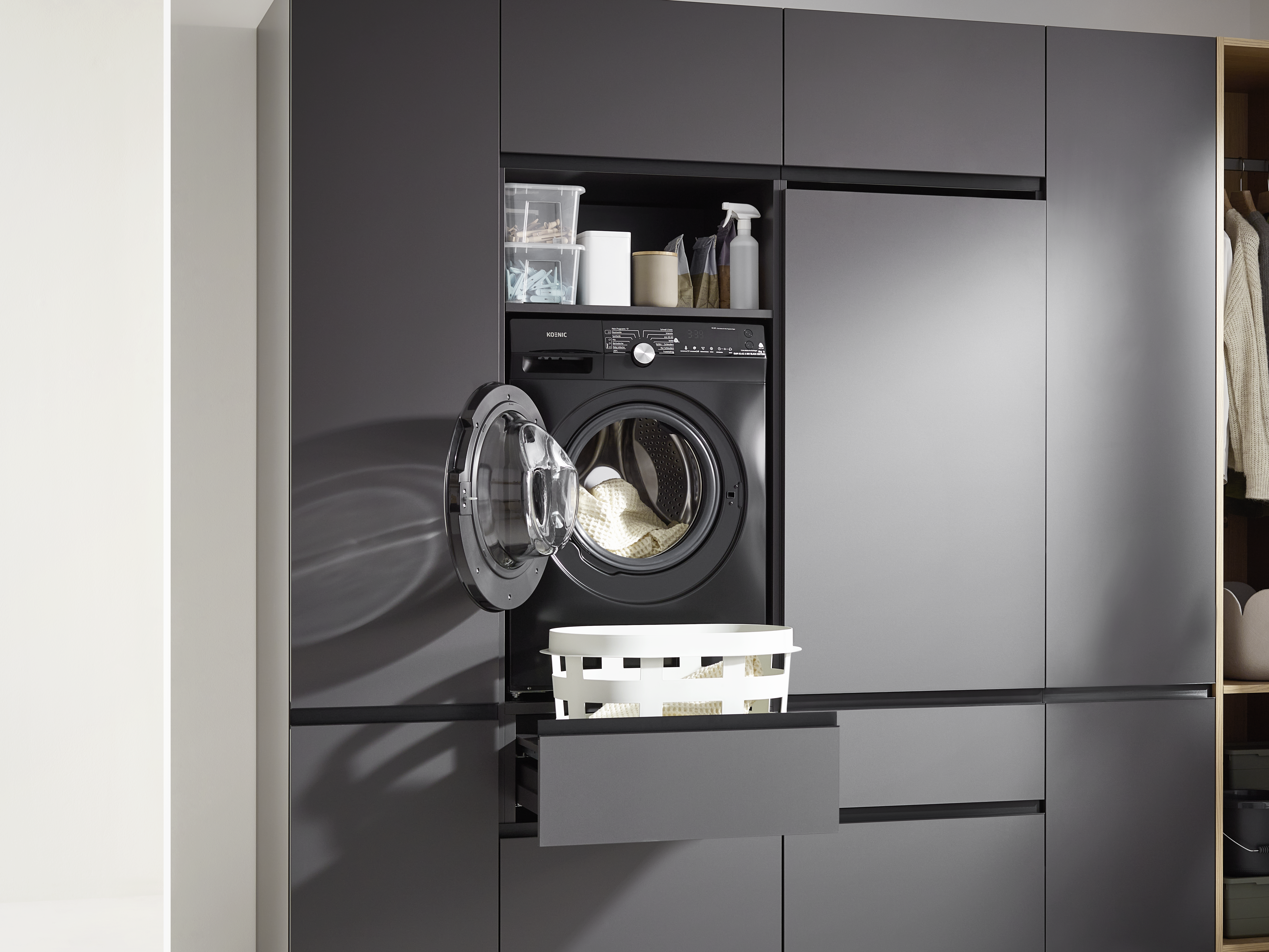 Eine schwarze Waschmaschine mit offener Tür, ein weißer Korb und Reinigungsmittel auf einem Regal. Der Hintergrund zeigt Schränke.