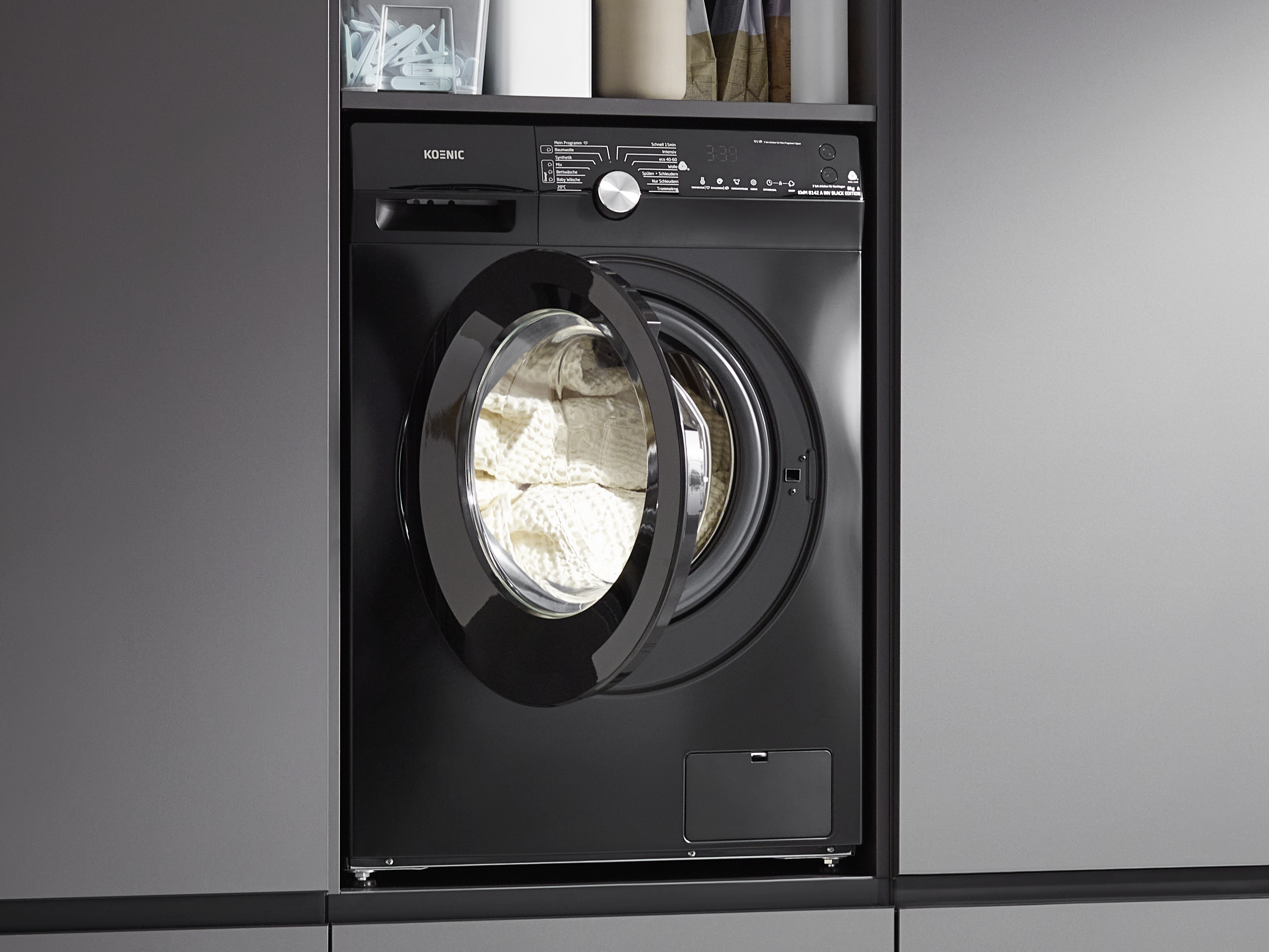 Schwarze Waschmaschine, offene Tür mit weißer Wäsche. Graue Schränke umgeben sie.