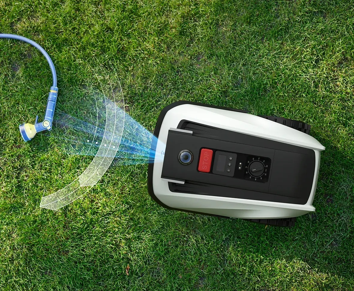 Robotyczna kosiarka do trawy na zielonej trawie. Wąż rozpyla wodę. Kosiarka ma czerwony przycisk stop.