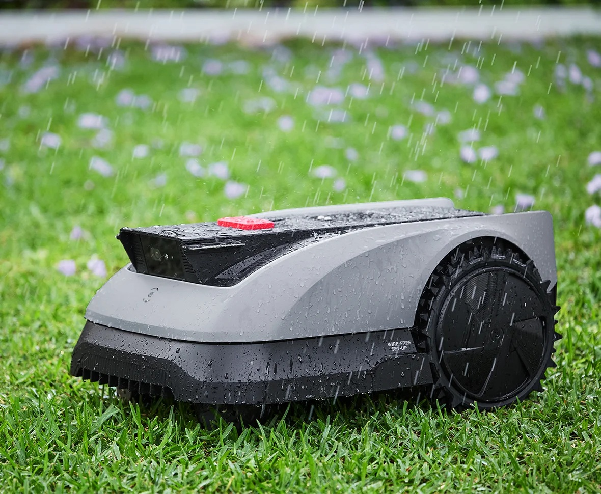 Robot koszący na zielonej trawie, w lekkim deszczu. Szary i czarny design, czerwony przycisk.