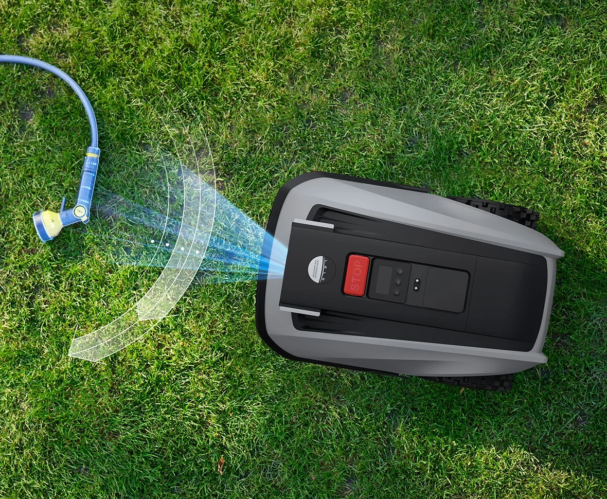 Kosiarka robot na trawie, z niebieskim wężem spryskującym wodą i widocznym przyciskiem stop.