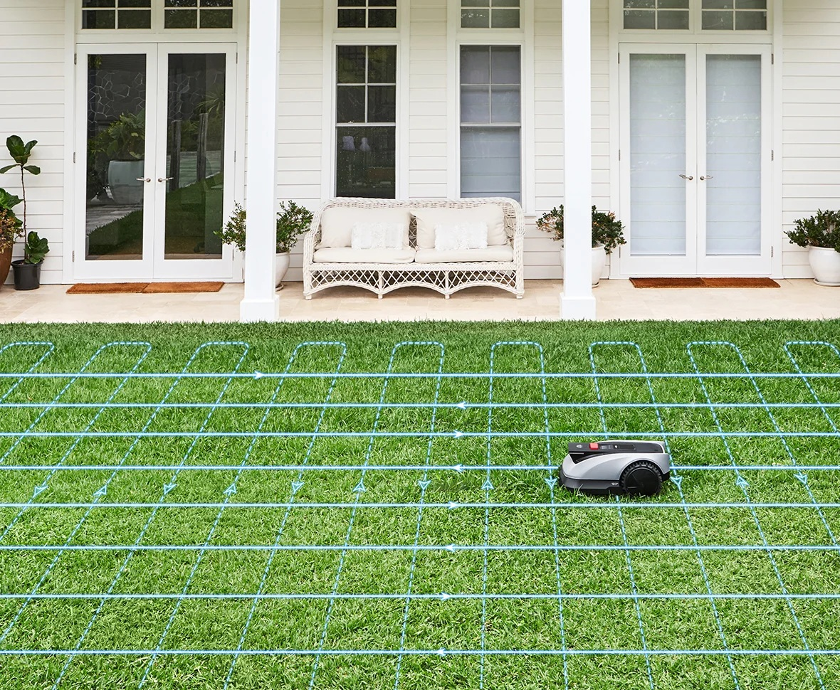 Kosiarka robot na zielonej trawie, z domem i niebieskimi liniami wyznaczającymi ścieżkę koszenia.