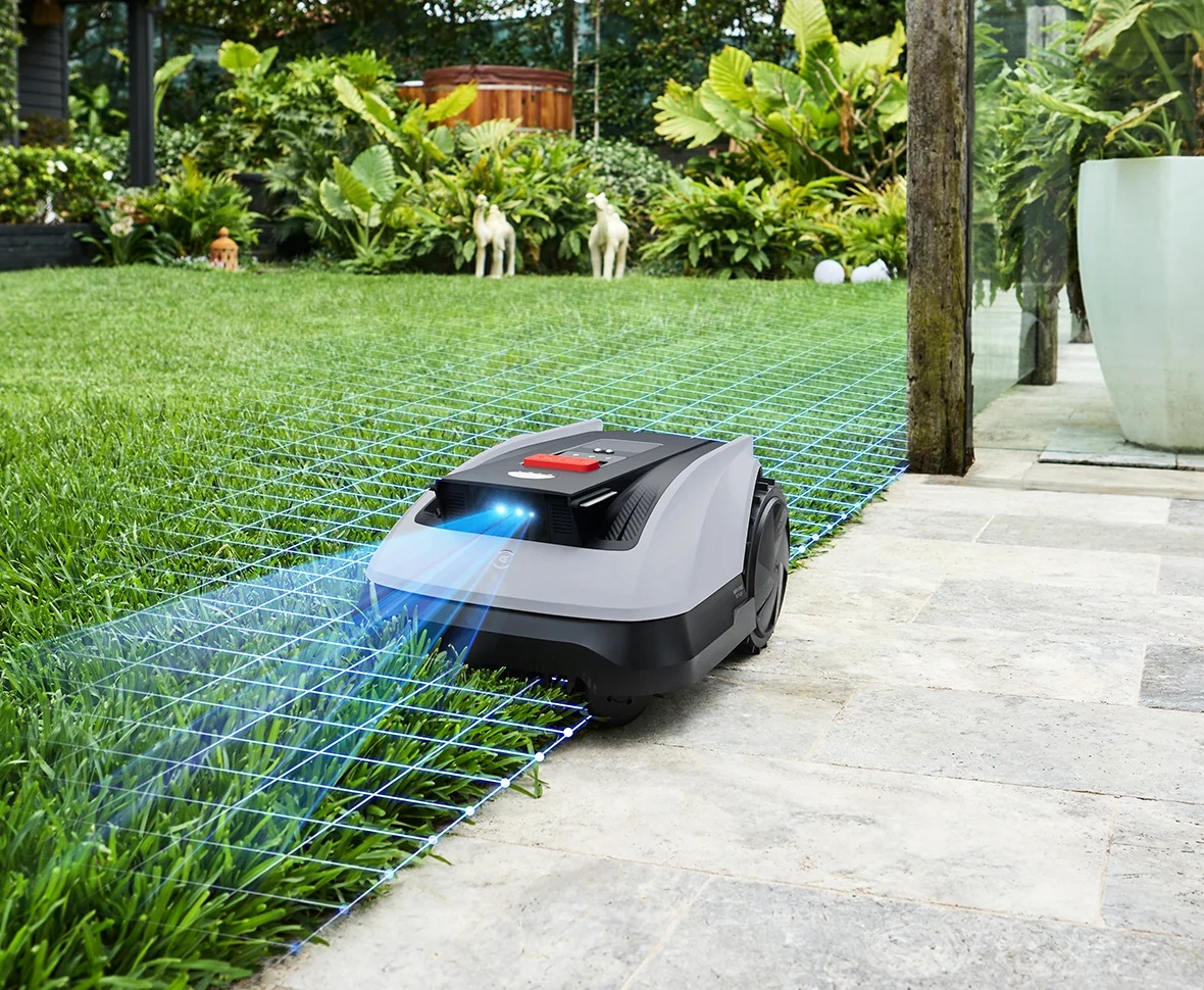 Robot koszący kosi trawnik. Niebieskie linie siatki. Zielona trawa, kamienna ścieżka, ogród z roślinami.