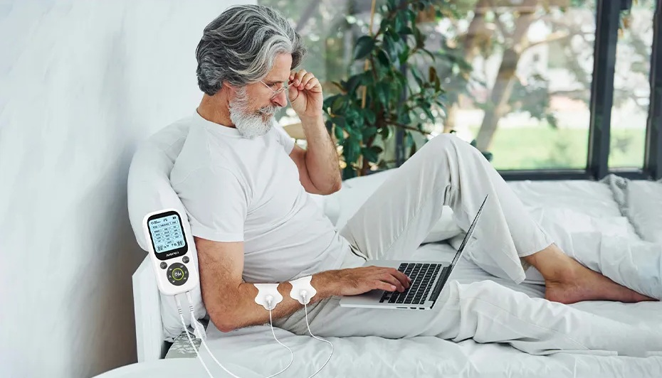 Mężczyzna siedzi na łóżku, używając laptopa, podłączony do urządzenia do stymulacji elektrycznej.