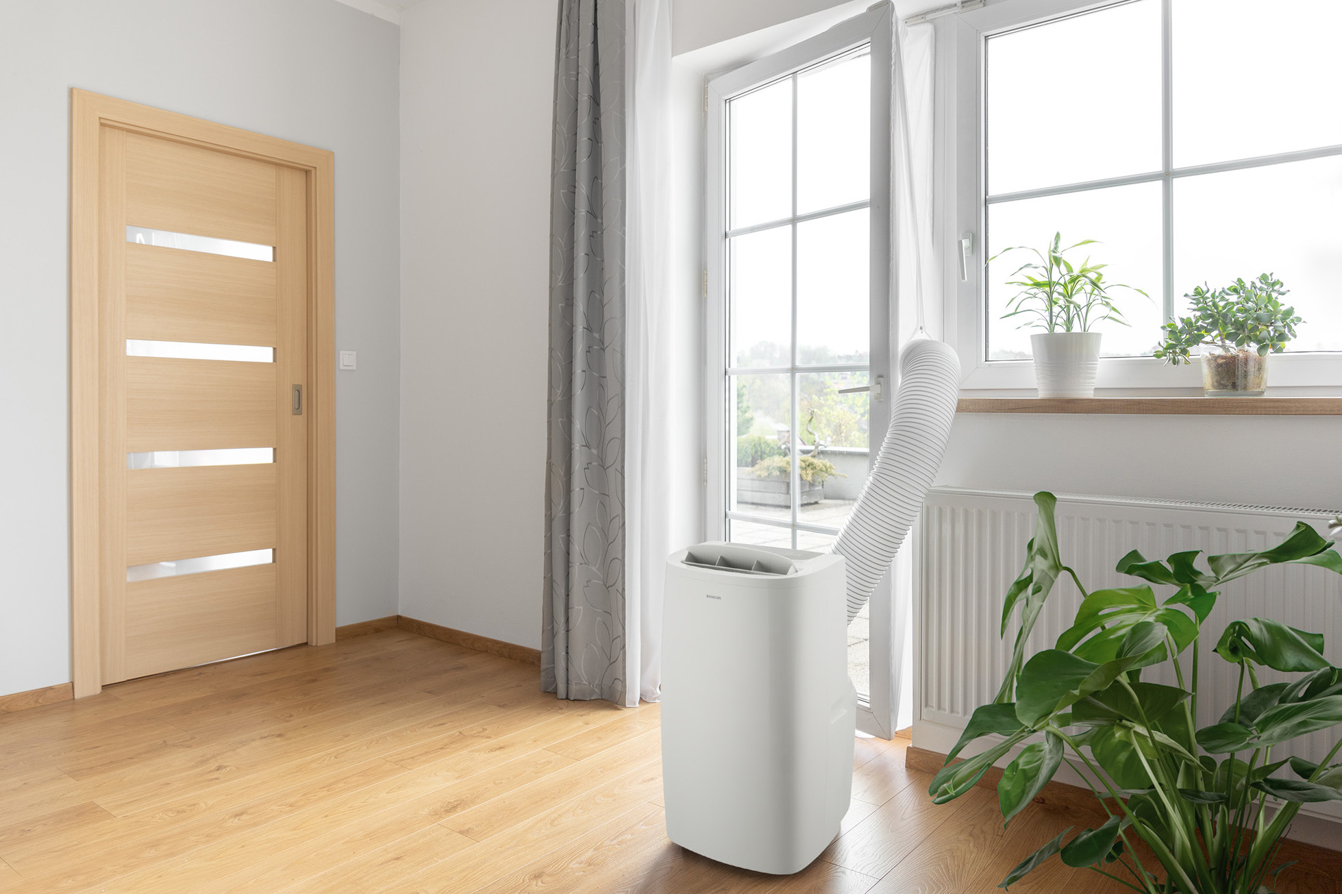Przenośny klimatyzator w pokoju. Widoczne drzwi, okno i rośliny. Białe urządzenie.