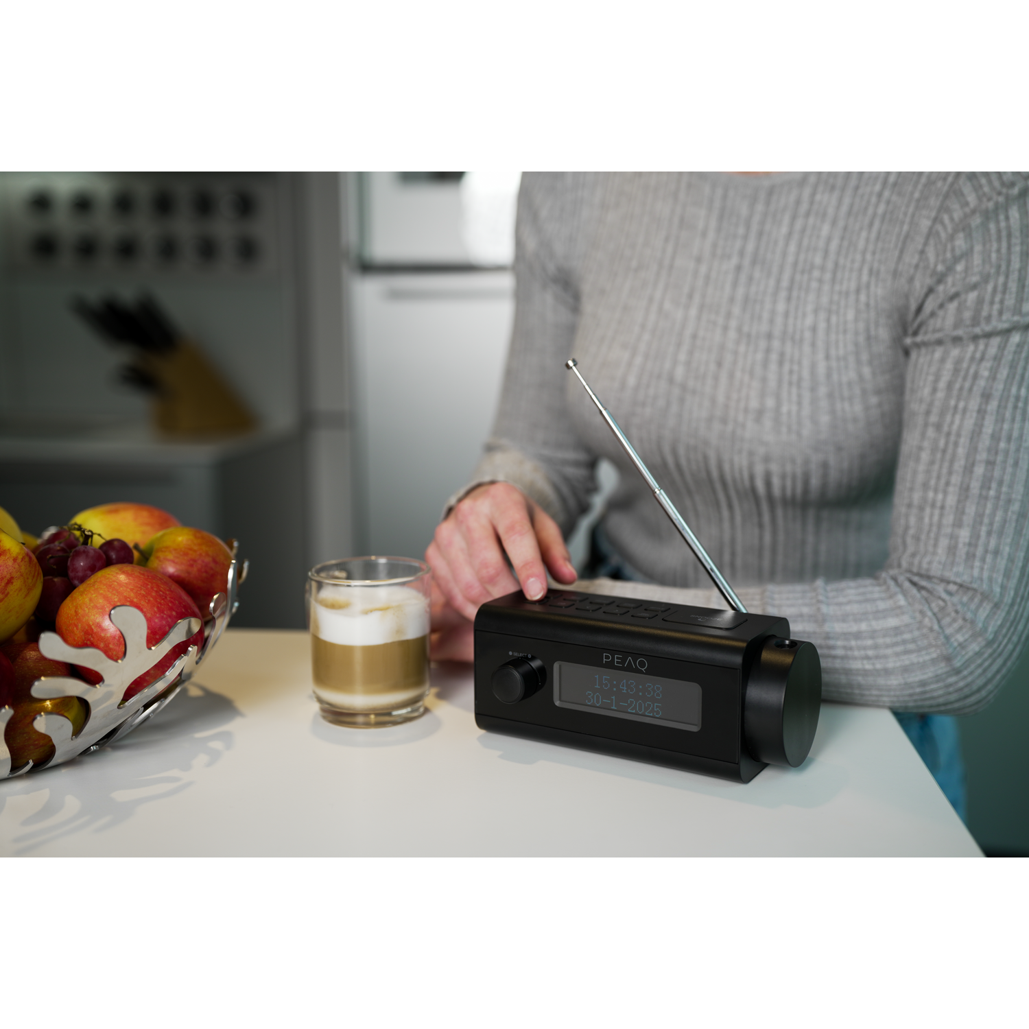 A person sits at a white table with a black radio, a glass of latte, and a bowl of fruit. The radio displays a date.