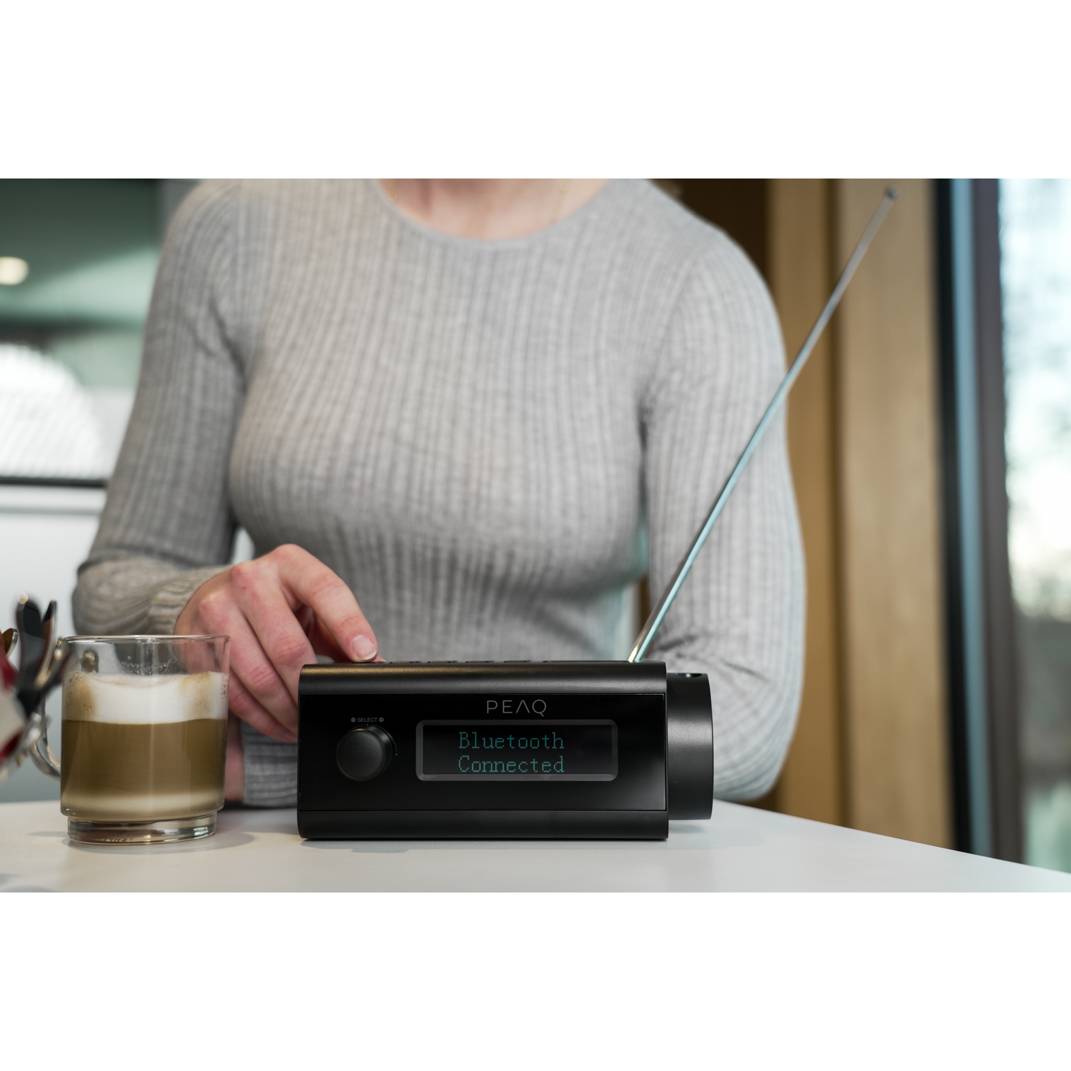 A woman is adjusting the volume of a black radio on a white table. A cup of coffee sits beside it.