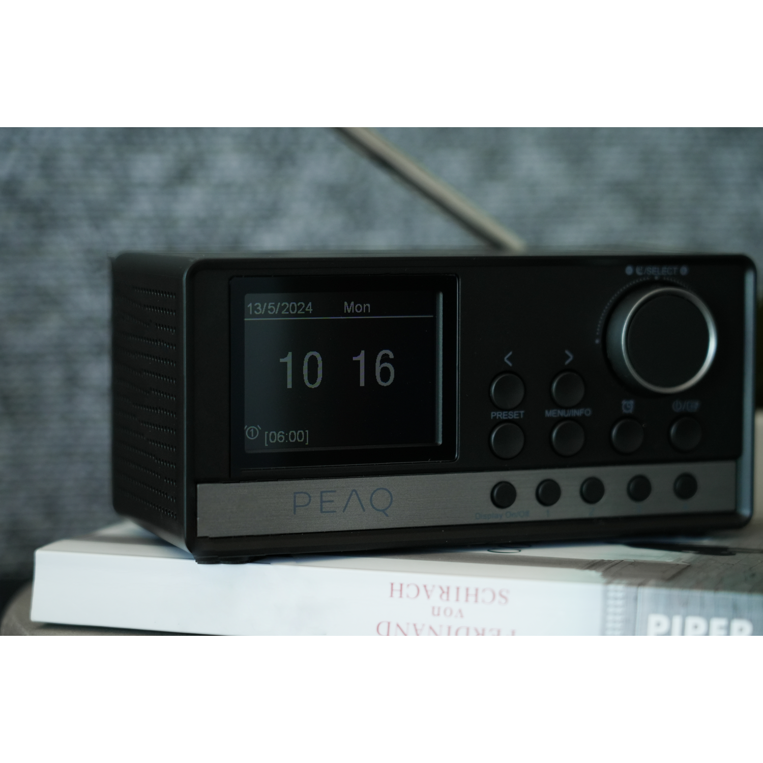Black radio with a screen showing time and date, buttons, and a dial. It sits on a book. The word PEAQ is on the front.