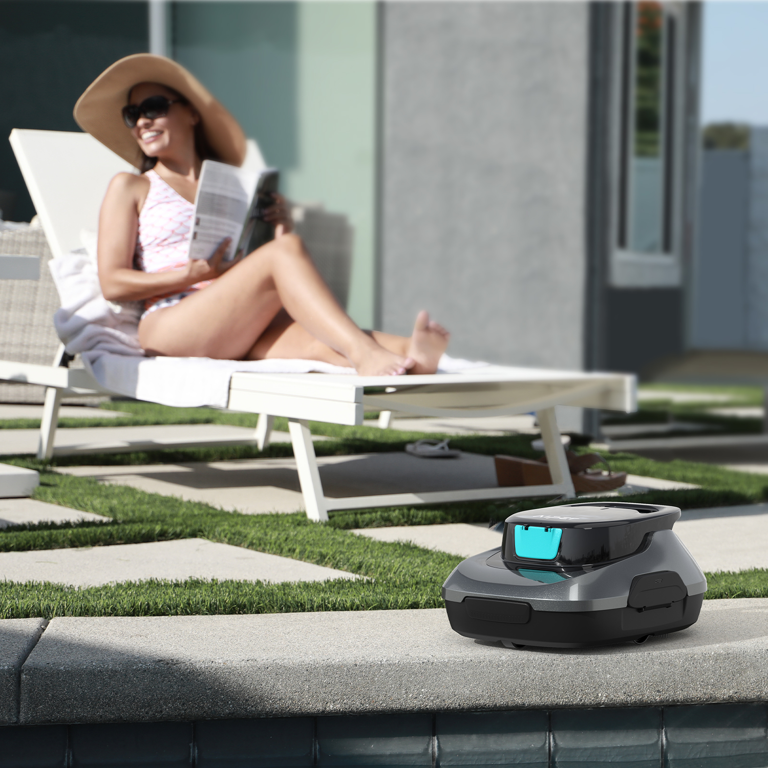 Woman reading, sunbathing near a pool. Robotic lawnmower on the pool edge in the foreground.