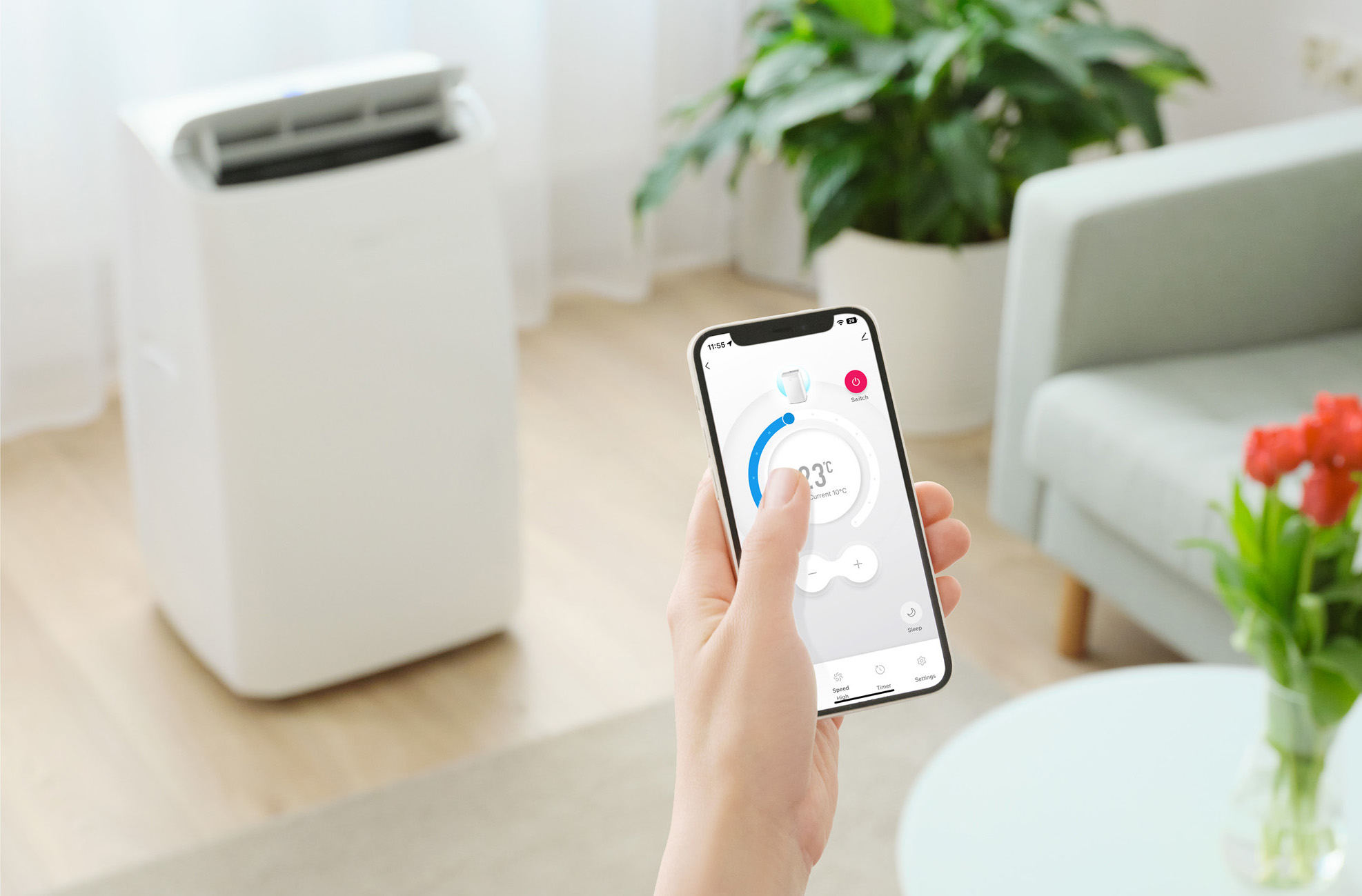 Osoba używa smartfona do sterowania białym oczyszczaczem powietrza. W tle pokój z roślinami i kwiatami.