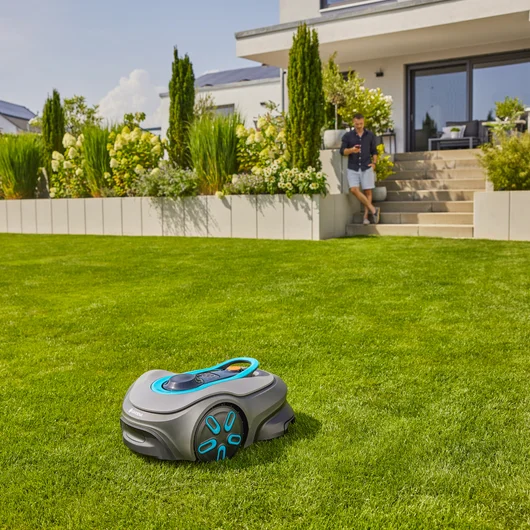 Mähroboter auf grünem Gras; Mann lehnt sich an Haus. Hintergrund: Garten, Haus und Himmel.