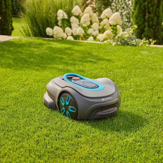Grauer Rasenmähroboter auf grünem Gras, mit blauem und orangefarbenem Bedienfeld. Weiße Blumen im Hintergrund.
