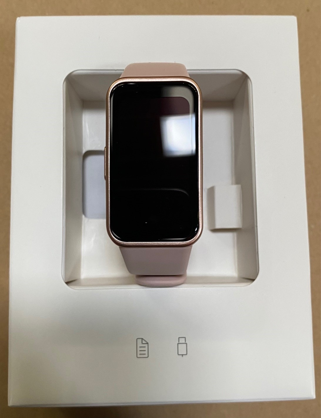 Smartwatch w białym pudełku. Zegarek ma różowy pasek i czarny ekran, ze złotą ramką.