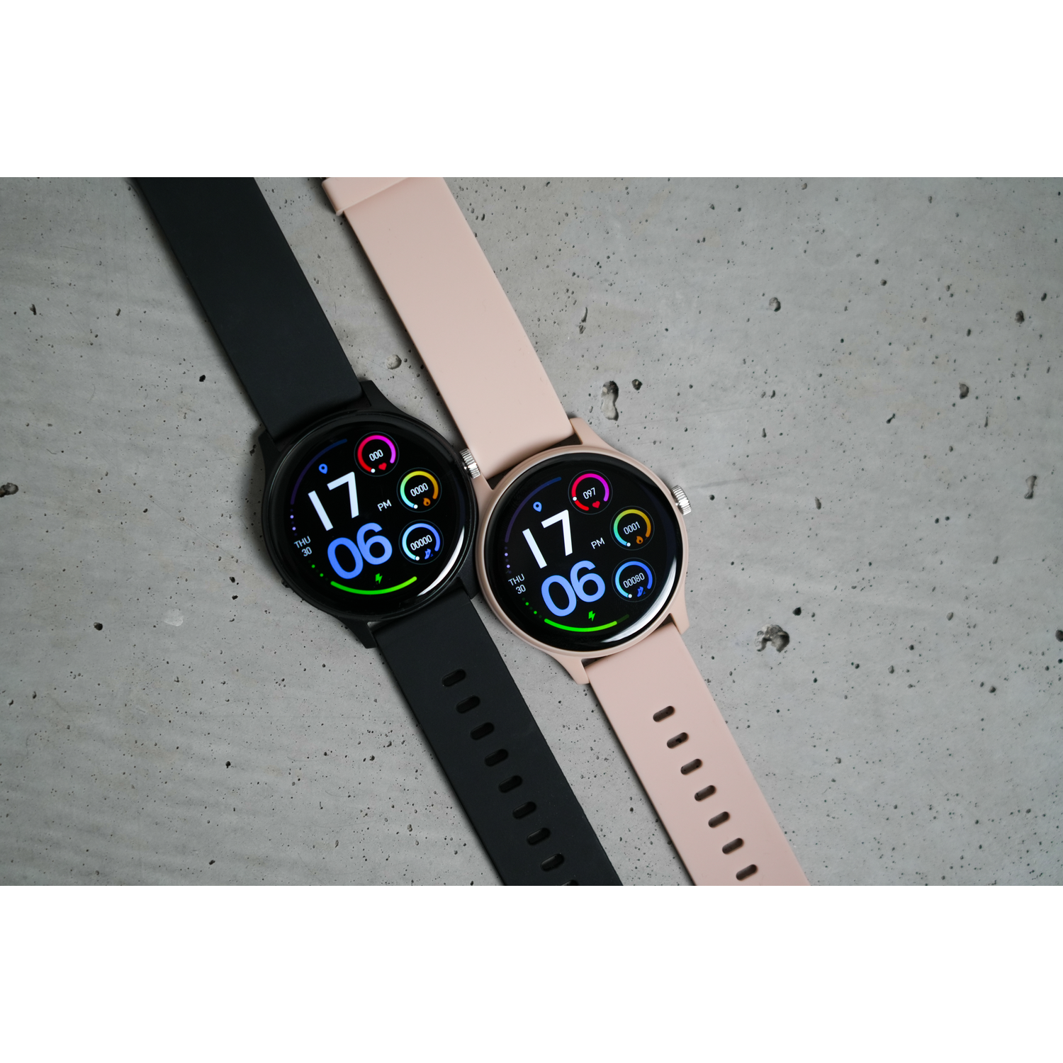 Two smartwatches, one black and one pink, displaying the date and time on a concrete surface.