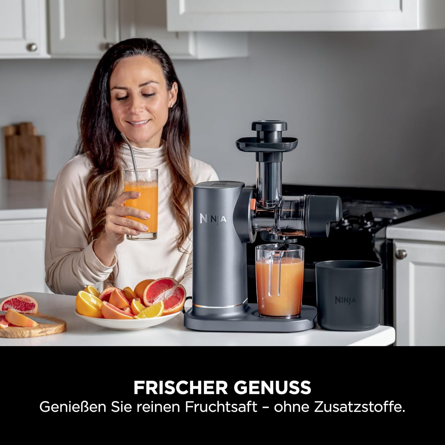 Una donna sorridente, che tiene in mano un bicchiere di succo. Una centrifuga e frutta in primo piano.
