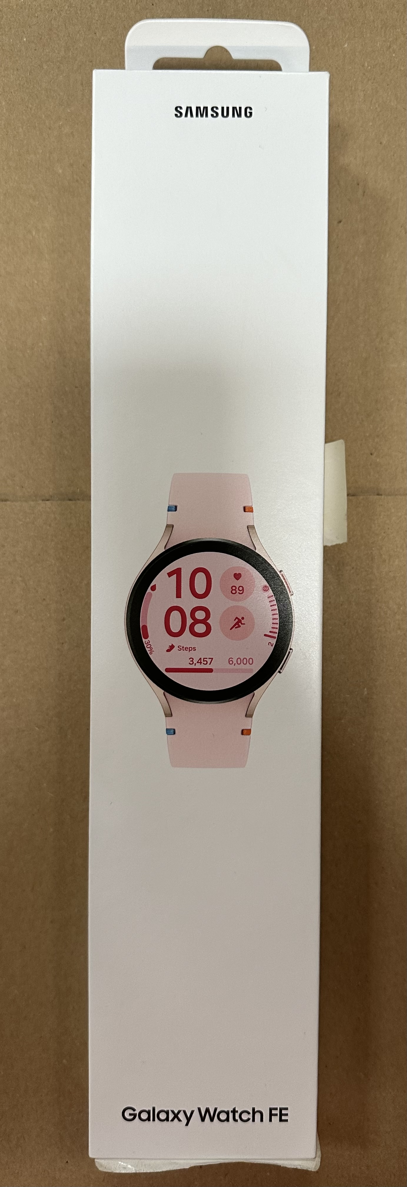 Różowy smartwatch, wyświetlający godzinę i inne dane, w białym pudełku z logo Samsung.