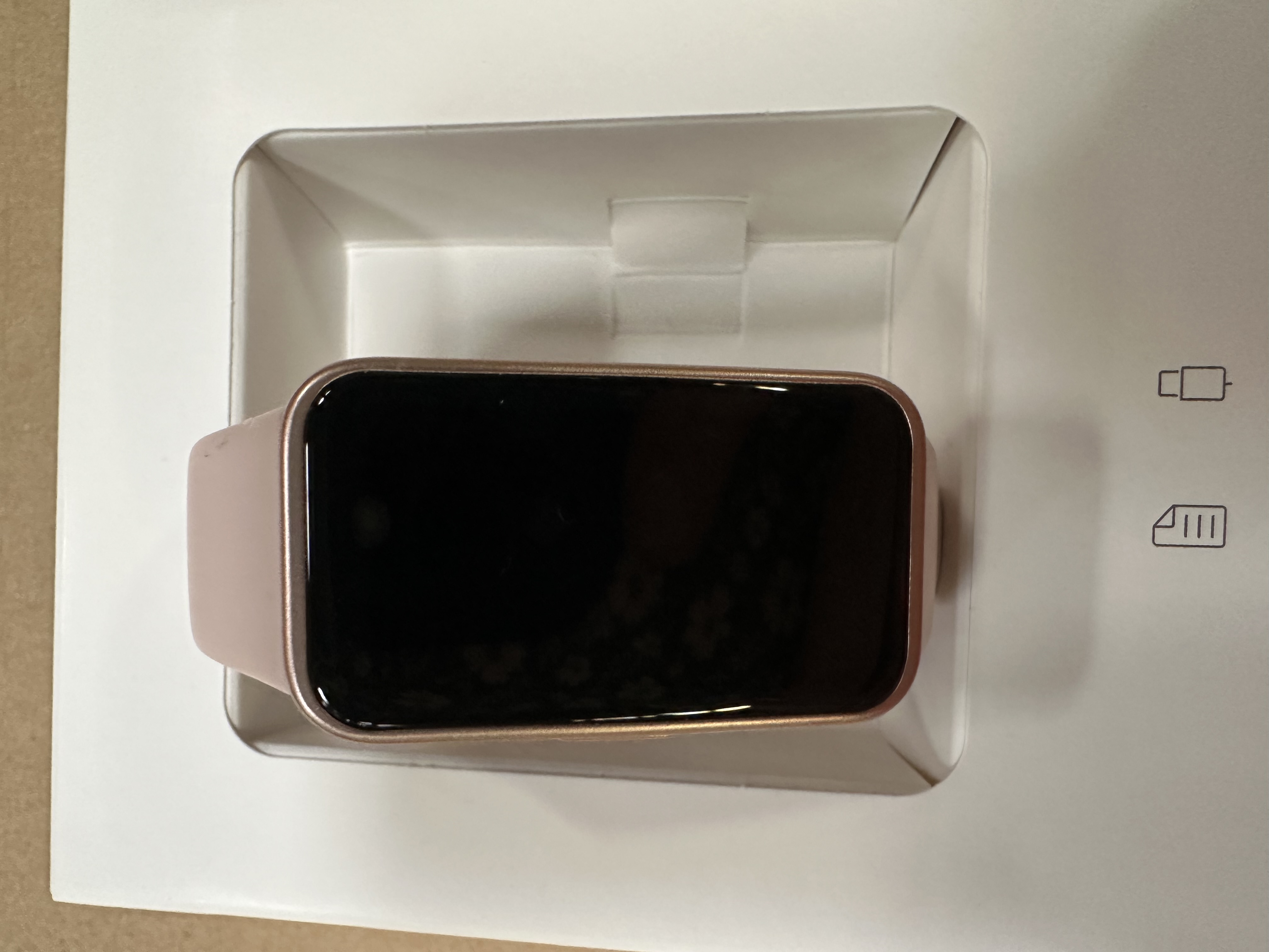 Różowy smartwatch w otwartym białym pudełku, z czarnym ekranem i kablem do ładowania.