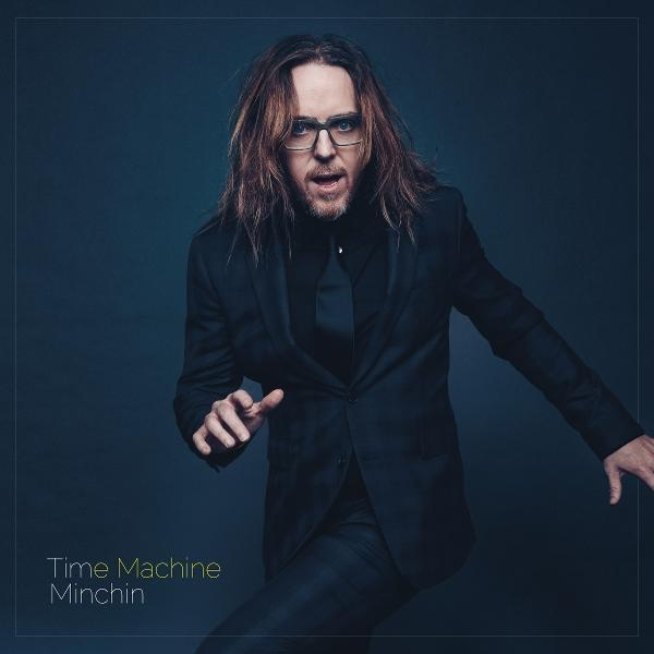 Mann in schwarzem Anzug, Brille und langen Haaren. Greift mit den Händen aus. Text unten: Time Machine Minchin.