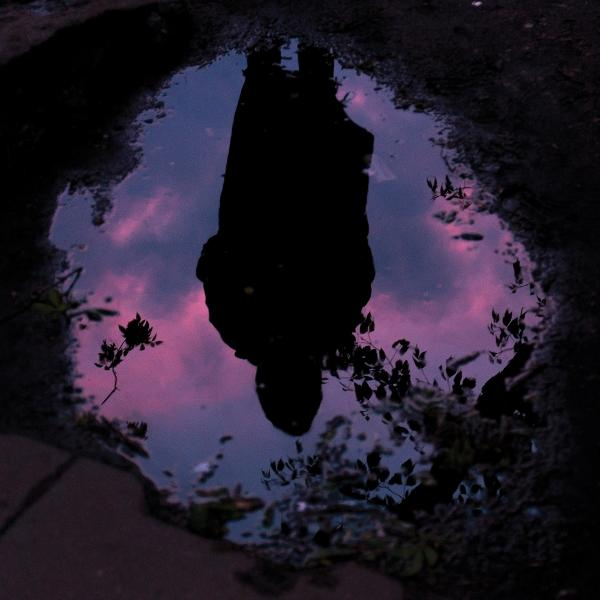Eine Pfütze, die einen lila-rosa Himmel und die Silhouette einer Person reflektiert.