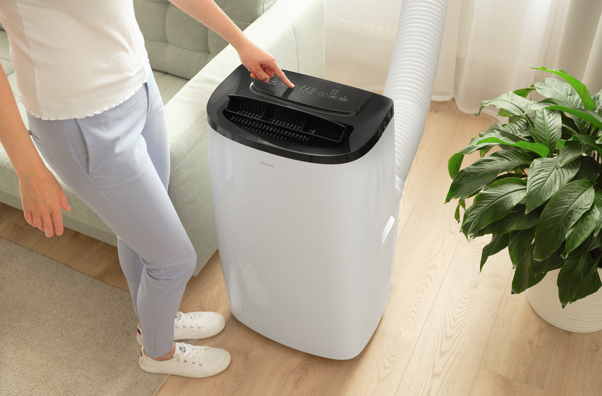 Osoba naciska przycisk na białym klimatyzatorze. Jest obok rośliny i sofy.
