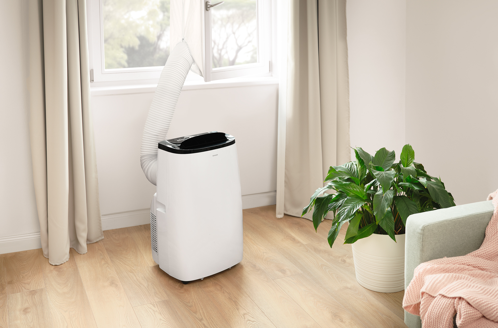 Biały przenośny klimatyzator z wężem. W pokoju z oknem, rośliną i zasłonami.