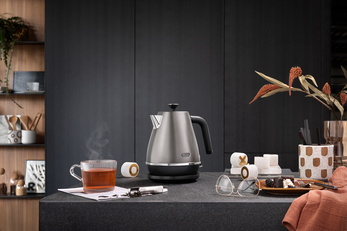 Srebrny czajnik DeLonghi na czarnej powierzchni, z herbatą i akcesoriami, ciemne tło.