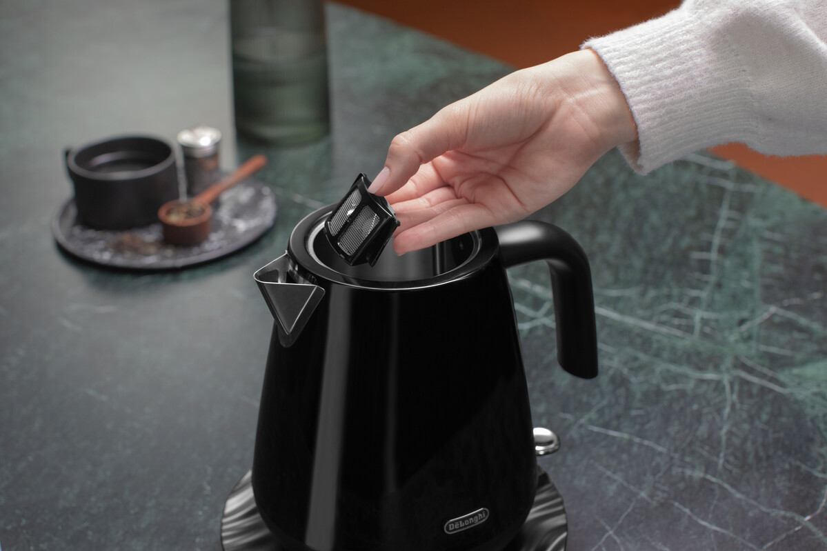 Ręka umieszcza filtr w czarnym czajniku DeLonghi. Czajnik stoi na marmurowym blacie.