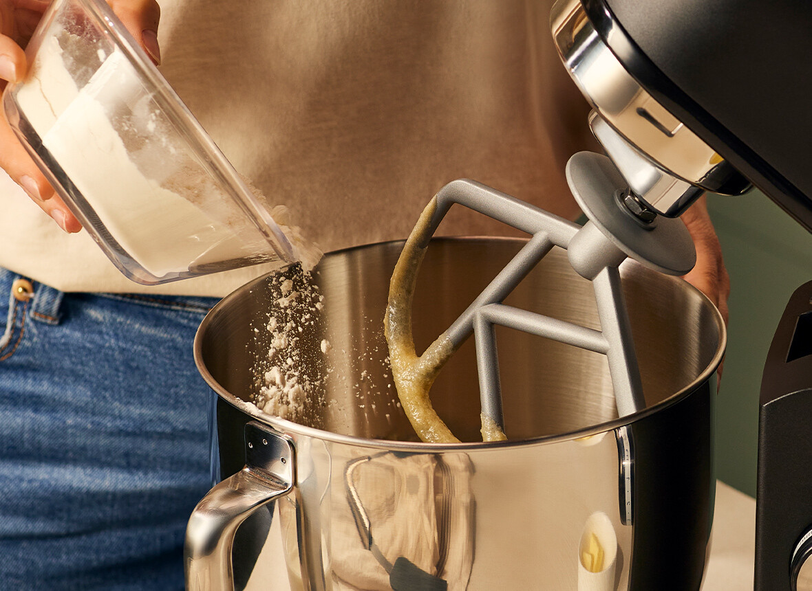 Dłonie wsypujące mąkę do miski z mikserem. Widok z bliska.