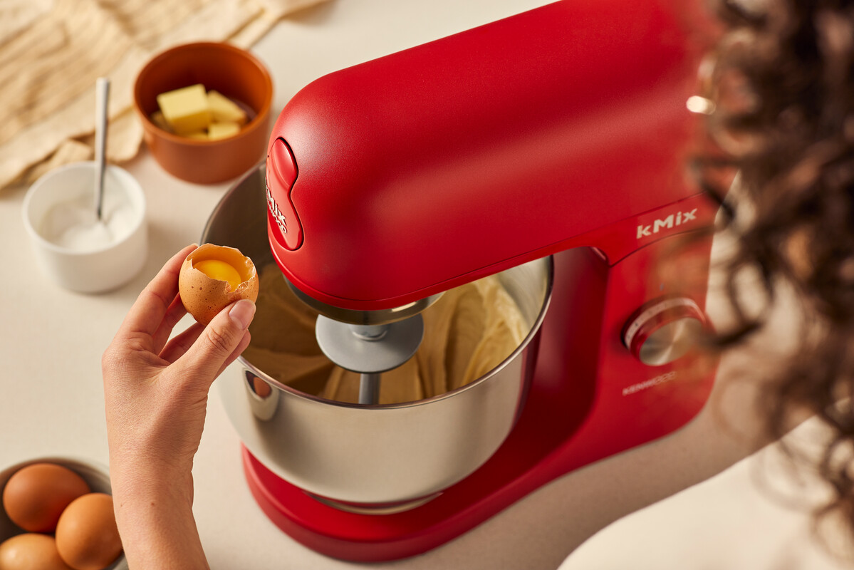 Czerwony mikser stojący z misą i jajkiem wbijanym do środka. Masło i cukier są na stole.
