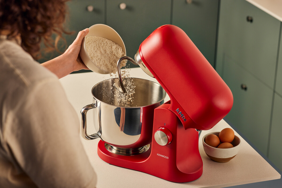 Osoba wsypuje mąkę do czerwonego miksera. Jajka obok w białej misce.
