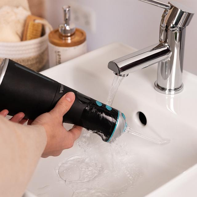 Osoba trzymająca czarny irygator nad białą umywalką z bieżącą wodą z chromowanej baterii.