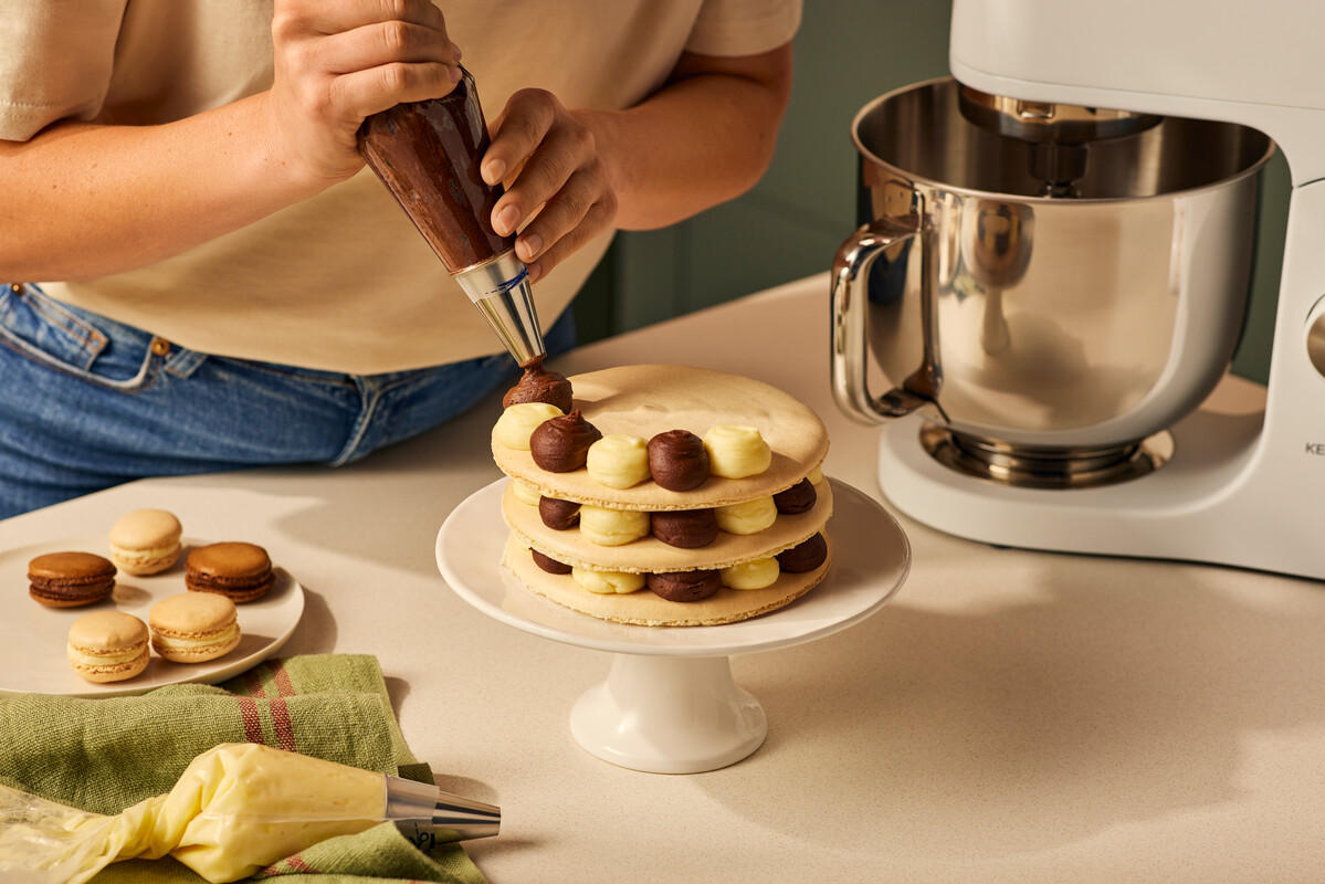 Osoba dekoruje ciasto lukrem. W tle makaroniki i mikser.
