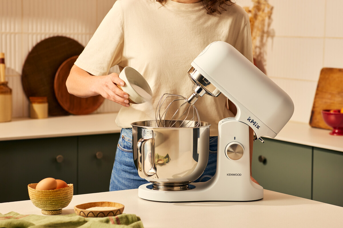 Kobieta dodaje cukier do białego miksera. Blat kuchenny z miskami, jajkami i składnikami.